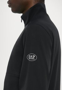 Juodas zip-up švarkas su aukštu apykakle, lygiu paviršiumi ir EA7 logotipu kairiajame rankove. Minimalistinio dizaino, be raštų.