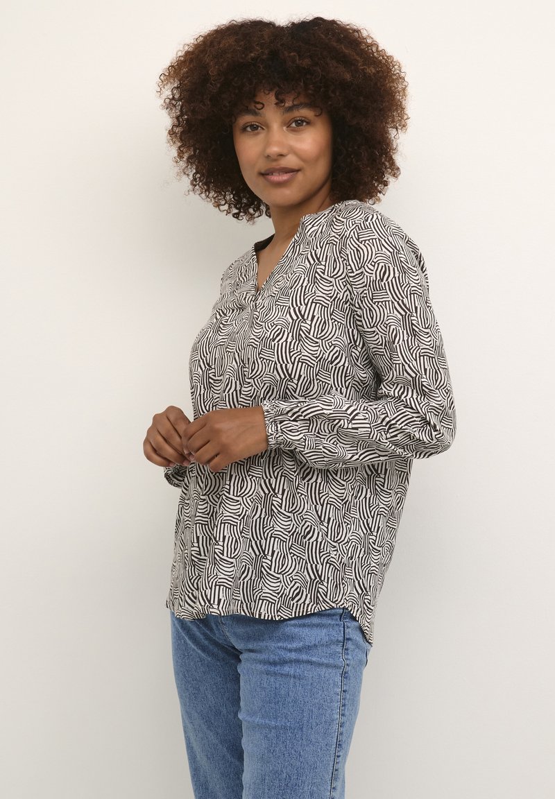 Langärmliges Oberteil in Schwarz und Weiß mit einem wellenförmigen Muster, V-Ausschnitt und lockerer Passform, kombiniert mit hellblauen Jeans.