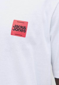 Hvid bomulds T-shirt med et rødt firkantet logo med teksten "Jack & Jones" i sort, placeret på venstre brystområde. Korte ærmer.