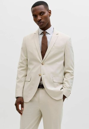 Mann trägt cremefarbenen Anzug, gestreiftes Hemd, braune Krawatte, steht vor schlichtem hellem Hintergrund mit einer Hand in der Tasche.