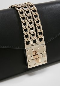 Sac à main en cuir noir avec des anses en chaîne dorée et un fermoir à tourniquet doré gravé "GUESS".