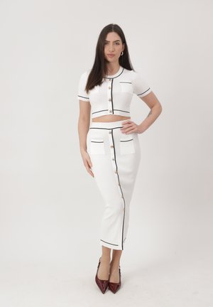 Vrouw met lang donker haar, gekleed in een wit, knoopjescrop top en bijpassende hoog getailleerde rok met zwarte biezen en bordeauxrode hakken, staand tegen een witte achtergrond.