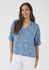 Svetlo modra kratka bluzica z diskretnim cvetličnim vzorcem, V-izrezom in sprednjimi gumbi. Narejena je iz teksturirane tkanine z sproščenim krojem.
