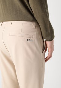 Beige pants made of smooth fabric, featuring a back pocket with a logo and side pockets, worn with an olive green long-sleeve top.
