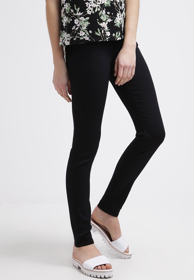 Femme portant un pantalon slim noir, un top fleuri et des sandales blanches à enfiler, debout avec une jambe légèrement pliée.