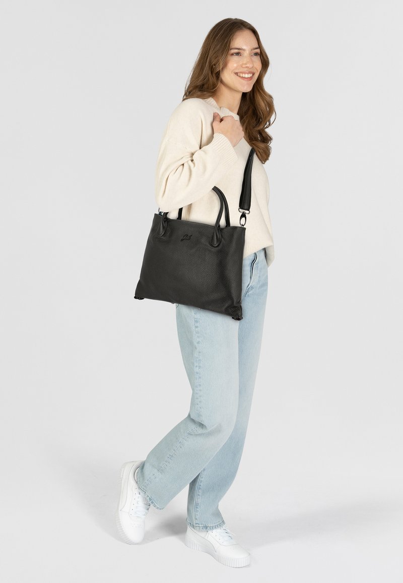 Bolso de mano de cuero negro con superficie texturizada, asas resistentes y correa ajustable. Modelo vistiendo un suéter claro y jeans azules.