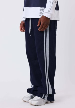 Homme portant un pantalon de survêtement bleu marine avec des bandes grises sur les côtés, des baskets blanches, et un t-shirt à manches longues superposé avec texte et motif d’étoile sur les manches.