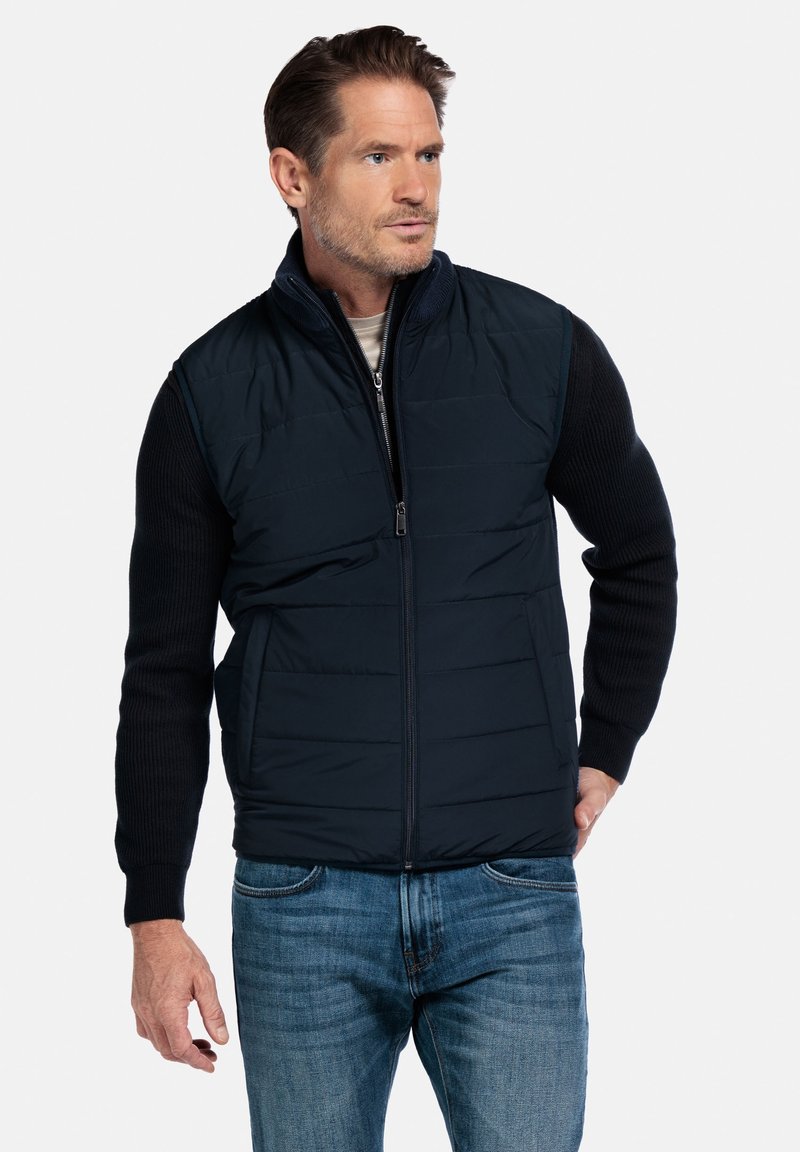 Marineblaue Steppweste mit hohem Kragen und Reißverschluss vorne, mit kontrastierenden, strukturierten Strickärmeln und zwei Seitentaschen. Über einem hellen Hemd getragen.