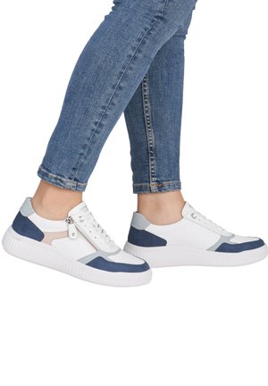 Person trägt weiße Sneaker mit blauen und grauen Akzenten und einem Seitenreißverschluss, kombiniert mit hochgekrempelten blauen Jeans.