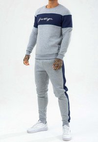 Szary sportowy sweatshirt z granatowym paskiem, okrągłym dekoltem i białym logo. Połączony z dopasowanymi dresami z granatowymi panelami po bokach i ściągaczami na rękawach.