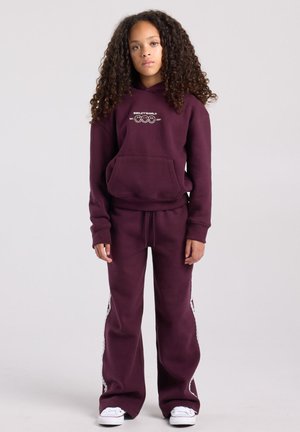 Jong persoon met krullend haar die een bordeauxrode hoodie en bijpassende joggingbroek draagt met witte sneakers, staand tegen een effen achtergrond.