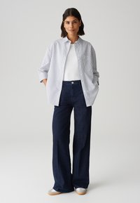 Camicia button-up a righe bianca e blu navy, abbinata a jeans a gamba larga blu scuro. Il modello indossa scarpe grigie con un design sottile.