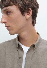 Light brown button-up shirt with a textured fabric, featuring a collar and a white undershirt visible beneath. Side profile of a person.