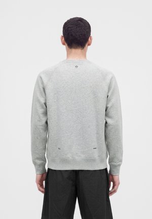 Uomo con felpa grigio chiaro a maniche lunghe e pantaloni neri, in piedi di spalle su uno sfondo chiaro uniforme.