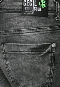 Jeans de mezclilla gris con un bolsillo trasero, remache de metal y una etiqueta negra con texto blanco y verde. Las texturas muestran un acabado desgastado.