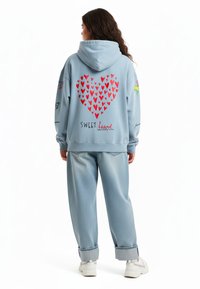 Modra kapuca s rdečim vzorcem srca, besedilom "SWEET heart" in različnimi pisanimi grafikami na rokavih. V kombinaciji z svetlo modrimi ohlapnimi džinsom.