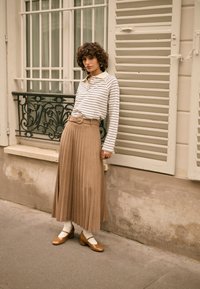 Bež plisirana suknja, bijela i tamnoplava prugasta majica dugih rukava, smeđe cipele s blok petom, bijele gležnjače, stoji kraj ukrasne ograde.