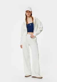 Witte hoodie en broek van zacht materiaal, gecombineerd met een marineblauwe crop top met een rood-wit grafisch ontwerp. Accessoires zijn een witte pet.