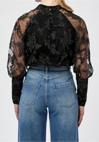 Blouse noire avec des manches sheer brodées de fleurs, associée à un jean en denim bleu taille haute. La vue de dos montre une fermeture à boutons et des poches soulignées.