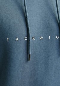 Sininen puuvillainen huppari, jossa on nyöri, ja jossa rintaosassa on valkoiset tekstit "JACK & J". Sileä kangas.