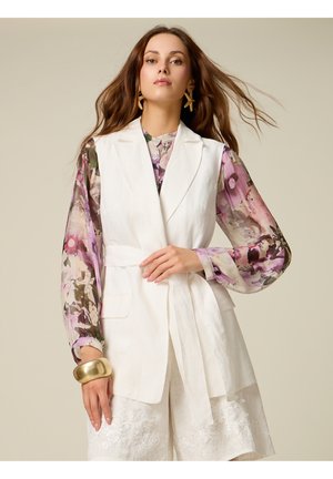 Mujer con cabello largo y castaño que lleva una blusa floral transparente, blazer blanco sin mangas con cinturón, pantalones cortos blancos de encaje, pulsera dorada y pendientes en forma de estrella.