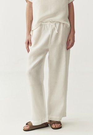 Pantalons en lin blancs avec une coupe décontractée, une taille à cordon de serrage et des jambes larges. Associés à des sandales à glissière brunes.