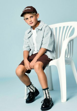 Enfant portant une chemise rayée à boutons, un t-shirt blanc, un short en velours côtelé marron et des chaussures noires avec des chaussettes rayées vertes et blanches, assis sur une chaise blanche.