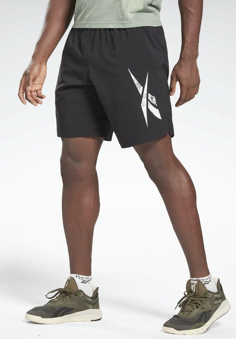 Persona atlética vestida con pantalones cortos negros de Reebok con logo blanco, zapatillas verde oliva de Reebok y calcetas blancas de marca, de pie sobre un fondo blanco.