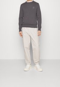 Mężczyzna ubrany w ciemnoszary sweter z długim rękawem, jasnobeżowe spodnie i białe trampki, stojący na tle jasnego, jednolitego tła.