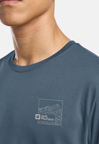 T-shirt blu scuro con collo rotondo, realizzata in tessuto morbido, con una grafica di montagna bianca e il logo "Jack Wolfskin" stampato sul petto.