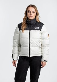 Chaqueta de plumas blanca con una sección superior negra y con patrones. Cuenta con un cierre de cremallera en la parte frontal, bolsillos laterales y acentos de logo en el pecho y la manga.