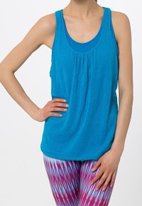 Kvinna iklädd en lös blå linne över en åtsittande blå sport-bh med mönstrade rosa och blå leggings, stående med en hand på höften.