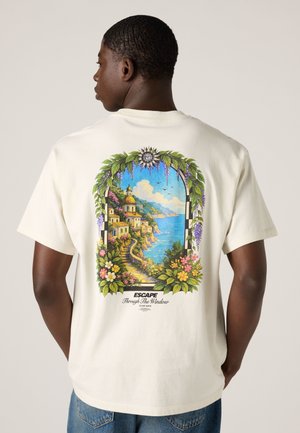 Hombre con camiseta blanca con un colorido pueblo costero y vista al océano enmarcados por flores y vegetación en la espalda.