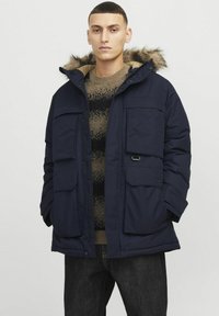 Marine parka jas met een faux fur-gevoerde capuchon, meerdere voorzakken en een gewatteerde textuur, die contrasteert met een bruine gebreide trui eronder.