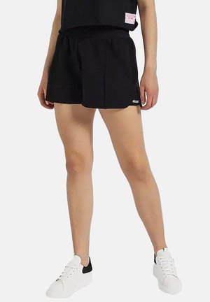 Personne portant un short décontracté noir et des baskets blanches, debout avec les mains détendues le long du corps devant un fond uni.