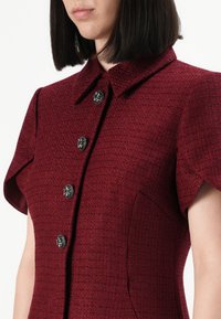Donna che indossa una giacca corta a maniche corte di un rosso profondo con una texture, dotata di colletto e quattro bottoni metallici decorativi lungo la parte anteriore.