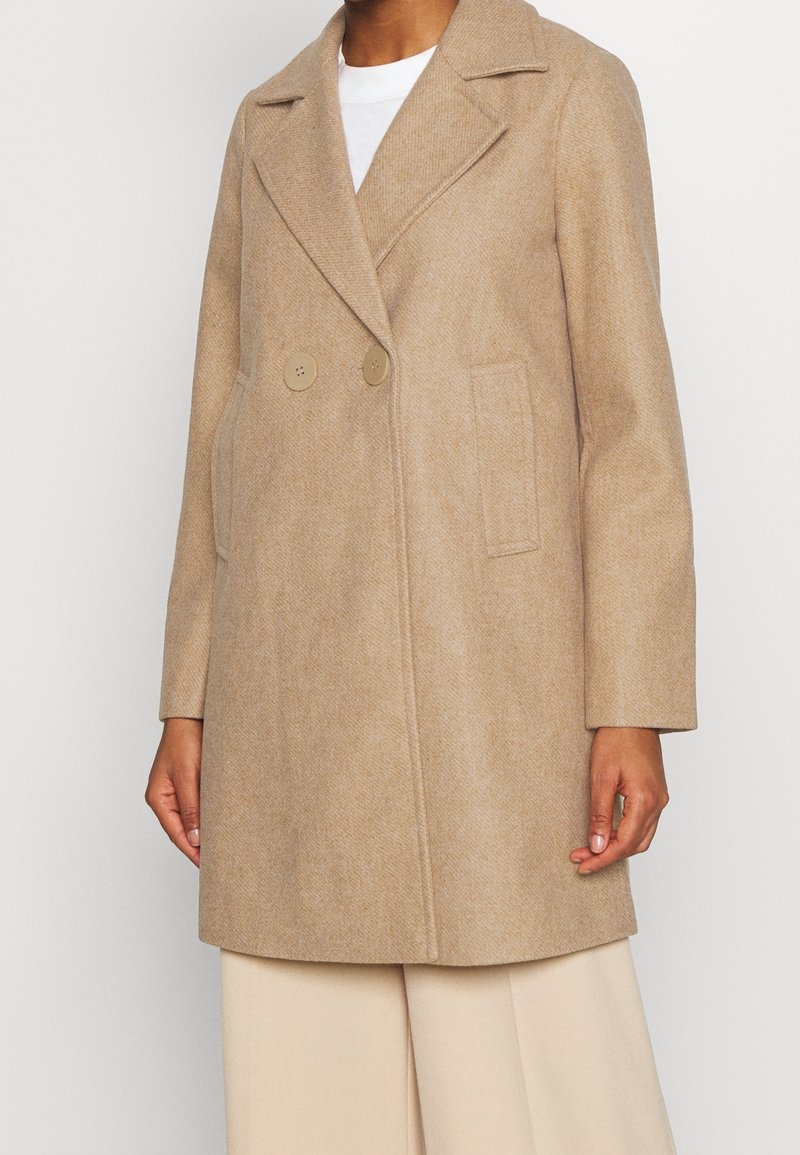 Abrigo beige de doble botonadura hecho de tela texturizada, con cuello pico, cierres de dos botones y bolsillos laterales.