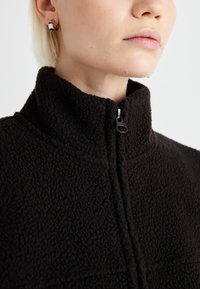 Schwarze Fleecejacke mit hohem Kragen, Reißverschluss und strukturierter Oberfläche. Hat eine enge Passform und ist aus warmem Material gefertigt.