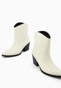 Białe skórzane botki za kostkę z wąskimi czubkami, dekoracyjnymi przeszyciami po bokach i czarnymi kwadratowymi obcasami. Gładka tekstura, nowoczesny zachodni design.