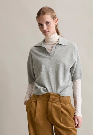 Grå kortermet genser med V-hals og krage, lagd over en hvit turtleneck. Brune kordfløyelsbukser med avslappet passform og legg.