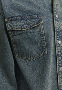 Denim-paita haaleassa sinisessä värissä, jossa on rintatasku, hopeiset nepparit ja strukturoitu kangas. Näkyvät saumat ja pehmeä laskeutuminen.