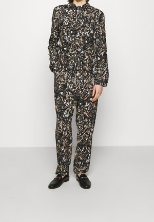 Henkilö pukeutuneena pitkähihainen eläinkuosisuunttuiseen haalariin, jossa on kaulus, ja mustat loaferit, seisoo yhdellä kädellä taskussa.