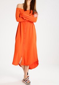Femme portant une robe orange vif à boutons et épaules dénudées, ainsi que des sandales à enfiler rayées, debout avec les bras croisés et les jambes croisées.