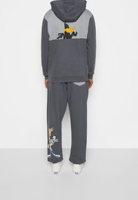 Sudadera con capucha gris con diseño de Pato Lucas en la parte posterior, combinada con pantalones grises que presentan un gráfico de Bugs Bunny. Ajuste casual y relajado.