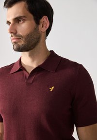 Bordeaux-polo paita, joka on valmistettu pehmeästä kankaasta, jossa on jämäkkä kaulus ja lyhyet hihat. Rinnassa on keltainen linnun emblem.
