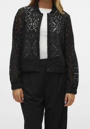 Femme porte une veste en dentelle noire boutonnée avec des motifs floraux sur un haut blanc et un pantalon noir sur un fond uni.
