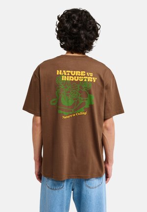 Osoba z kręconymi włosami, ubrana w brązowy t-shirt z zielonym motywem roślinnym oraz żółtym napisem „Nature vs Industry” i „Nature is Calling!” na plecach.