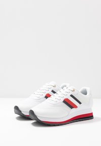 Baskets de sport blanches avec des rayures latérales noires, blanches et rouges, lacets blancs et accents de semelle rouges et noirs, présentées côte à côte sur fond blanc.