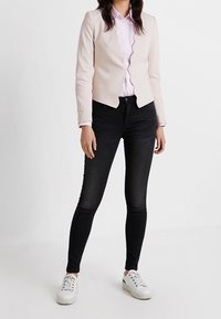 Blazer ajusté rose clair avec bords festonnés, porté sur une chemise violet clair, associé à un jean skinny noir et des baskets blanches.