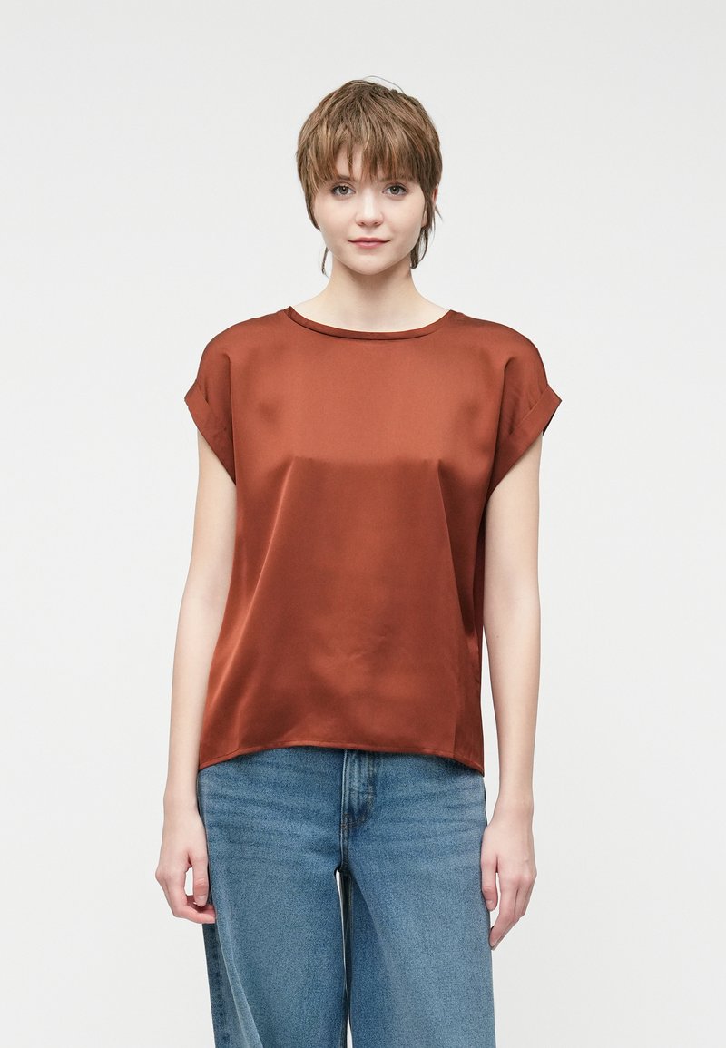 Ruda šilko bluzė su apvaliu kaklu, trumpomis rankovėmis ir laisvu siluetu, derinama su šviesiai mėlynomis džinsinėmis kelnėmis iki juosmens.